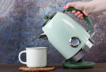 A woman pours hot water from a green electric kettle into a light-colored cup.