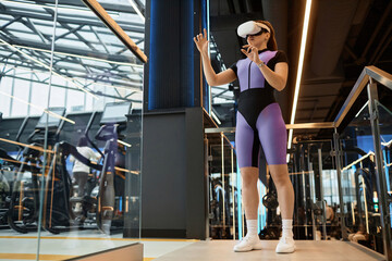 Full length portrait of sportive young woman wearing VR headset in gym and using interactive...