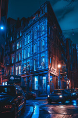 Naklejka premium Moody evening setting featuring a striking building bathed in blue light on a street corner with wet roads reflecting the ambiance