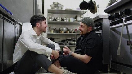 A cheerful guy a brunette waiter in a white shirt communicates with his colleague a cook in a black uniform during a break at work in a restaurant in the kitchen