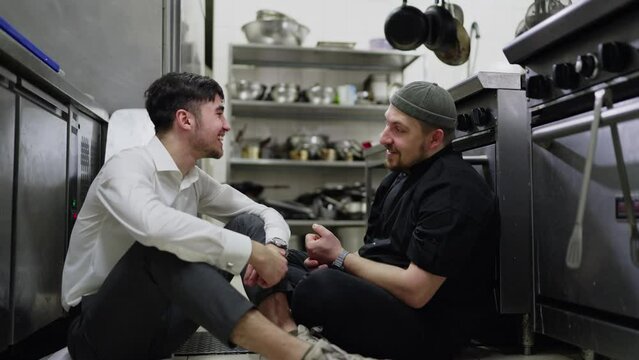 A cheerful guy a brunette waiter in a white shirt communicates with his colleague a cook in a black uniform during a break at work in a restaurant in the kitchen