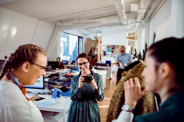 Smiling female fashion designer holding a camera in a lively design studio with colleagues examining fabric in the background