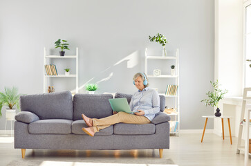 Elderly gray-haired senior woman relaxing using laptop sitting on couch in the living room at home in headphones. E-learning for mature people or distance freelance work for retirement.
