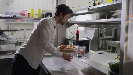 A confident brunette male waiter in a shirt takes a dish and delivers it to the hall taking it from the serving table in the kitchen in a restaurant. Interaction between waiters and cooks when issuing