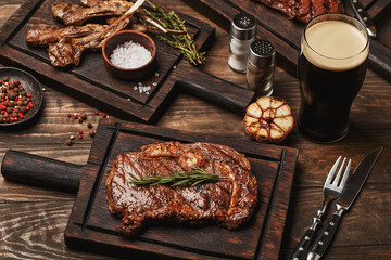 Wooden table served with various grilled meat, vegetables and glass of beer. Striploin steak, ribeye steak and lamb ribs on wooden cutting boards
