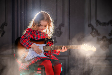 little girl holds a guitar and sits against the curtain in the rays of the spotlights.