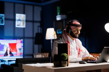 Smiling Arab man with wireless headphones, typing and doing research on his personal computer. Male...