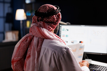 Focused Arab businessman diligently works at his desk, conducting online tasks and examining...
