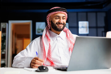 Smiling Arab guy works diligently at office desk, engages in digital tasks and writes on his...