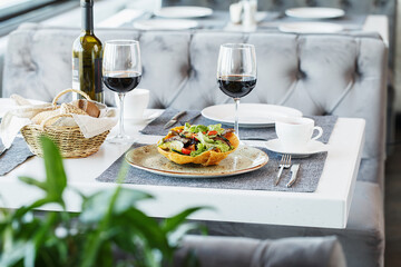 Restaurant table served with tableware, salad, glasses and bottle of red wine and bread basket. Fine dining. Table setting. Restaurant interior