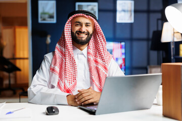 Male person dressed in traditional Arabic clothing is seated with his laptop. Confident and...