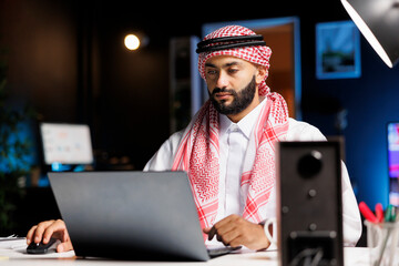 Arab businessman with confidence typing on a laptop at a contemporary desk. Male Muslim person using his personal computer for email and browsing in a serene and professional workplace environment.