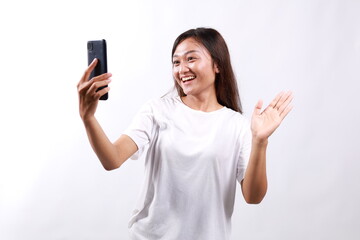 Portrait of friendly young asian woman talking on video chat app, waving hand at smartphone camera, having mobile conversation, standing against white background