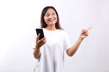 Excited young asian woman shopping on mobile app, holding phone and pointing finger left at empty copy space, showing promo text, standing with smartphone against white background