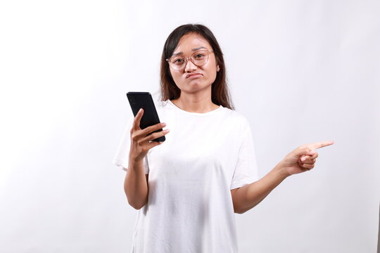 Skeptical asian modern girl, holding smartphone, pointing finger left at empty space and frowing hesitant, has doubts. Isolated on white background