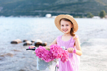 Kid with bike on sea beach. Child girl in straw hat riding on sunny coastline. Pink hydrangea...