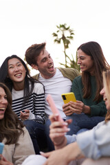 Vertical. Group diverse people enjoying together outdoors with mobiles in hand. Young multiracial friends using cell phone. Beautiful millennial sitting together laughing excited at class break