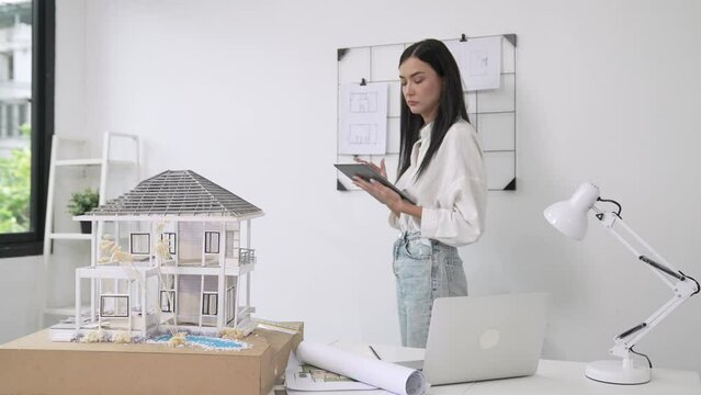 Beautiful caucasian architect focus on using tablet to check house model on meeting table with blueprint, house model and architectural document scatter around at meeting room. Immaculate.