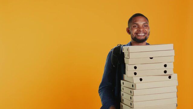 Male pizzeria courier carrying huge pile of pizza boxes in studio, preparing to deliver meal order to customers. Young deliveryman holding a big stack of fast food takeaway, shipping. Camera B.