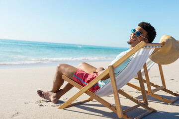 At beach, young biracial man relaxing on beach chair, enjoying sun, copy space