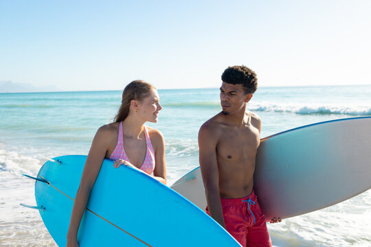 At beach, diverse couple holding surfboards, looking at each other