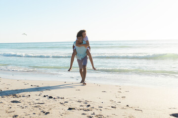 At beach, diverse couple enjoying piggyback ride, laughing together