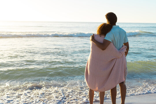 At beach, diverse couple embracing, watching waves