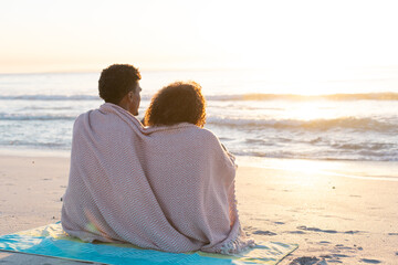 Obraz premium At beach, diverse couple sitting close under blanket, watching sunset