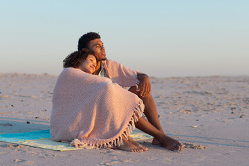 Obraz premium At beach, biracial couple wrapped in blanket, gazing at horizon