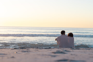 Obraz premium At beach, diverse couple sitting close, watching waves, copy space
