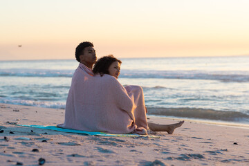 Obraz premium At beach, biracial couple wrapped in blanket, gazing at sea
