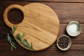 Cutting board, salt, pepper and rosemary on wooden table, flat lay. Space for text