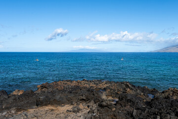 Couple of people stand up paddleboarding in the Pacific Ocean off the black lava rock point on...