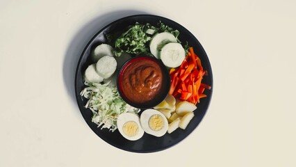 Gado-gado served on a plate, isolated white, Indonesian salad containing boiled vegetables and potatoes, boiled eggs, fried tofu tempeh, lontong and melinjo chips, served with peanut-cashew sauce. 