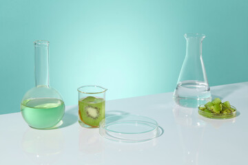 Baby blue background with gradient effect against white table contains some experiment utensils with a blank petri dish for showing product and fresh kiwi. Blank space for design with high angle shot
