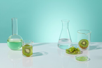 White countertop containing a boiling flask with light green liquid, fresh kiwi in beaker and chopped kiwi in petri dish, vacant space in center for displaying. Front view photo, copy space