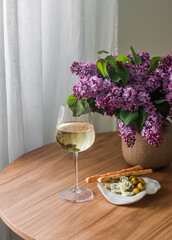 Delicious aperitif - a glass of white wine, a plate with snacks, a bouquet of lilacs on a wooden table