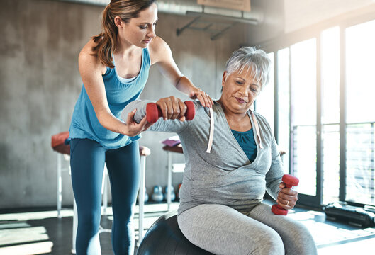 Senior woman, weights and helping with coach in gym for physio, arm exercise and rehabilitation on ball. Old female patient, instructor and workout equipment for arthritis with balance and dumbbells