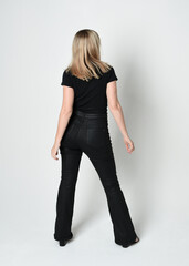 Full length portrait of beautiful blonde woman wearing modern black shirt and leather pants. Confident  standing pose walking away from camera, silhouetted on white studio background.