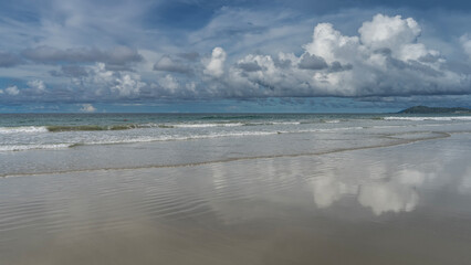 The waves of the endless turquoise ocean roll towards the shore, foaming and spreading along the beach. Clouds in the blue sky. Reflection on the wet smooth sand. Malaysia. Borneo. Kota Kinabalu