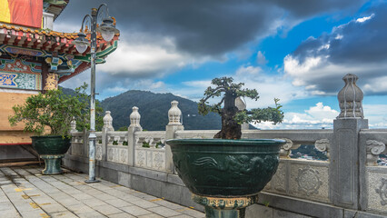Dwarf bonsai trees grow in pots on the terrace of a Chinese temple. The walls and curved roof are decorated with ornaments. A street lamp. In the distance, against a blue sky and clouds -a green hill.