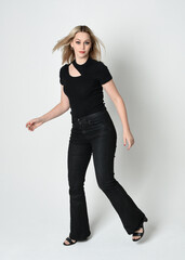 Fototapeta premium Full length portrait of beautiful blonde woman wearing modern black shirt and leather pants. Confident standing pose with gestural hands presenting, silhouetted on white studio background.