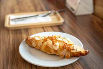 A piece of baked croissant with cheese and almond topping in a white plate and placed on wooden...