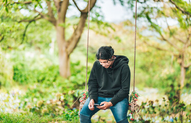 Smiling guy sitting on a swing with smart phone. Young man sitting on a swing using cell phone in nature