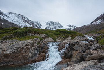 Gloomy alpine landscape with mountain creek in green valley among snowy mountains in rainy weather. Beautiful waterfall on cold river among big rocks and stones near grassy hill under gray cloudy sky.