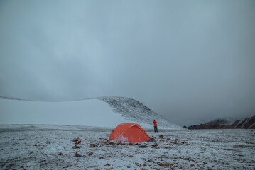 Man in red jacket in misty high mountains in freshly fallen snow. Orange tent on snow-covered stony...