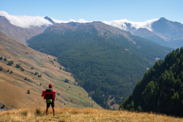 Fototapeta premium Young man hiking with backpack, staring out to green valley with mountains in the background