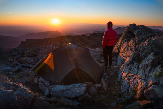 young man standing next to tent on a mountain at sunrise - Powered by Adobe