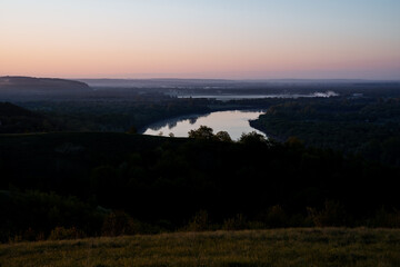 A serene lake is embraced by trees on a hill under the sunset sky