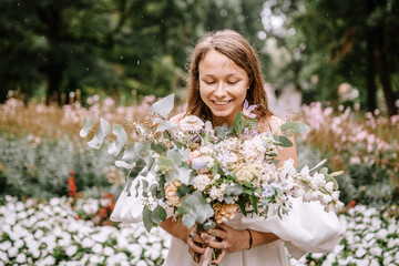 Riga, Latvia, - August 26, 2024 - Bride smiling joyfully while holding a bouquet, surrounded by a garden with white flowers.